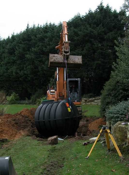 Pelleteuse posant une fosse toutes eaux en polyéthylène dans une fouille, au fond d'un jardin